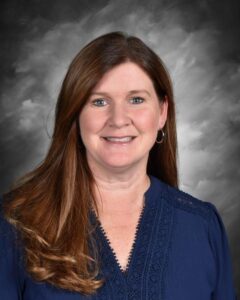 A smiling woman with long brown hair wearing a blue blouse, set against a gray background.
