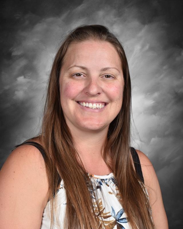 A smiling woman with long brown hair, wearing a floral top, against a gray background.