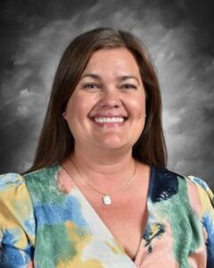 Smiling woman with long brown hair, wearing a colorful blouse, against a gray backdrop.