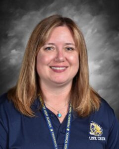 Smiling woman with long brown hair in a navy shirt, wearing a necklace, against a gray background.