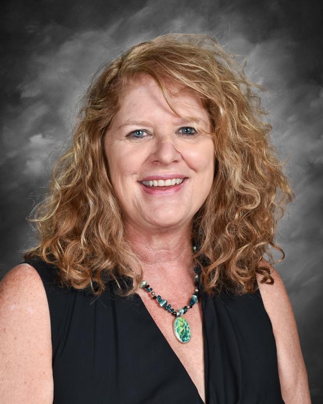 Smiling person with curly hair, wearing a black top and a turquoise necklace, against a gray background.