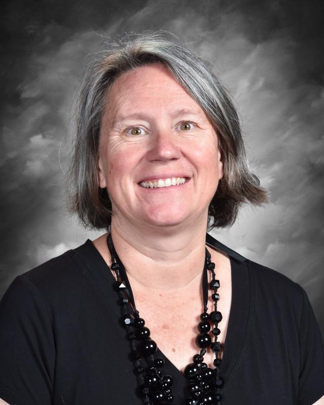 Smiling woman with short gray hair wearing a black top and beaded necklace, set against a gray background.
