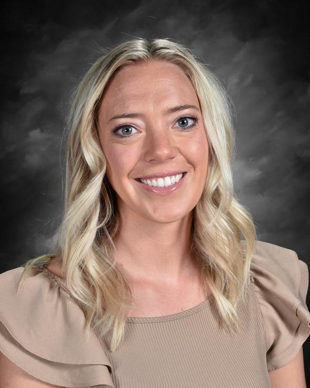 A smiling woman with long, wavy blonde hair, wearing a beige top, against a dark, textured background.
