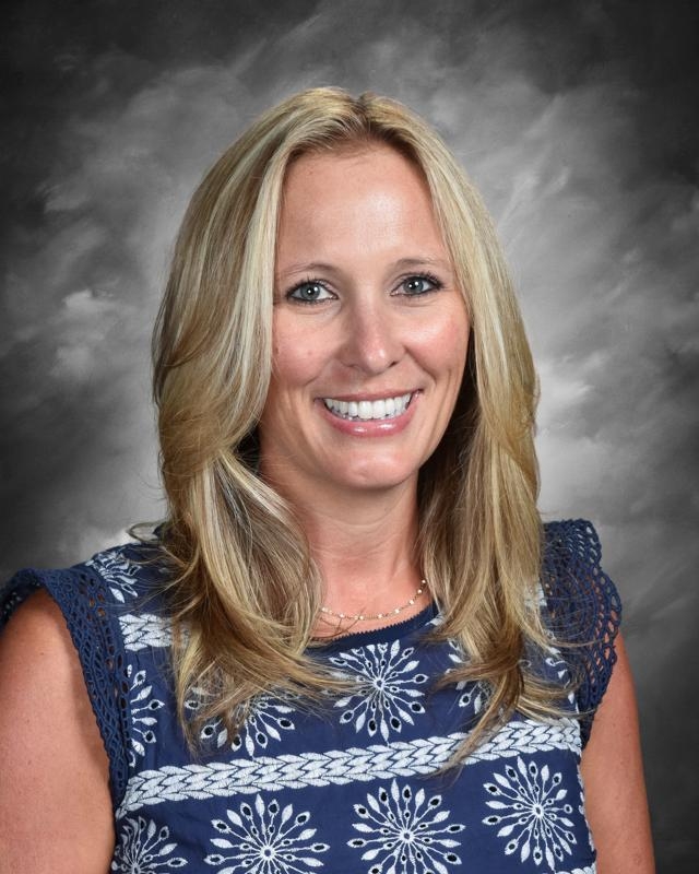 Smiling woman with long blonde hair in a blue patterned top, set against a gray background.