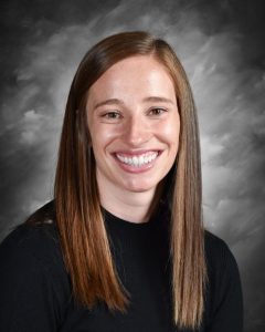Smiling person with long brown hair wearing a black top, set against a gray background.