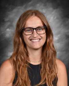 A smiling woman with long, wavy red hair and glasses, wearing a black top, against a gray background.