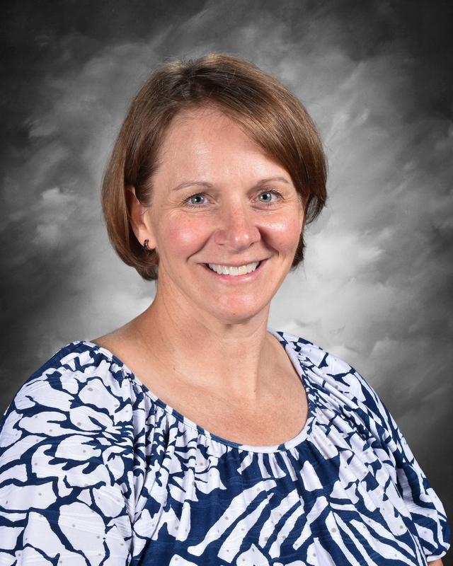 A woman with short brown hair smiles, wearing a blue and white patterned shirt, against a gray background.