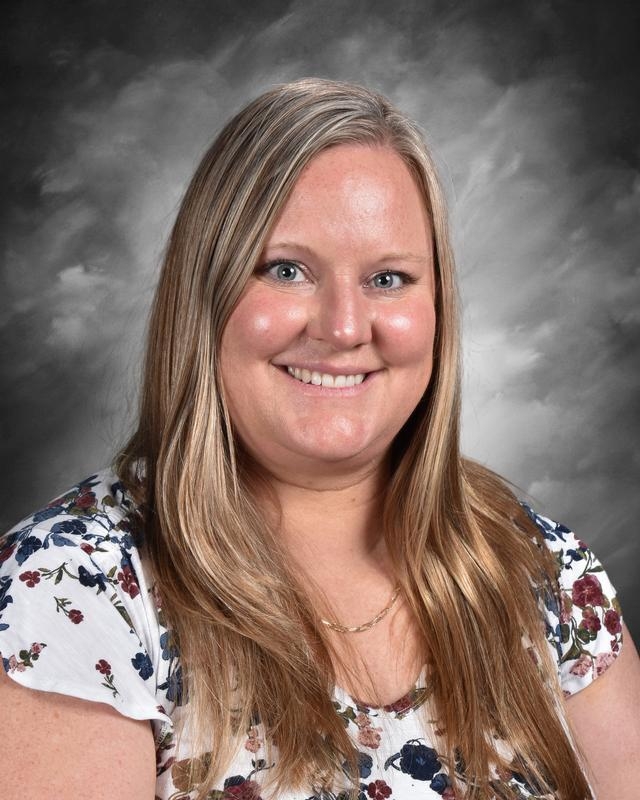 A woman with long blonde hair smiles, wearing a floral blouse, against a gray background.