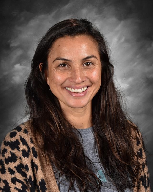 Smiling woman with long dark hair, wearing a gray shirt and a leopard-print cardigan against a gray background.