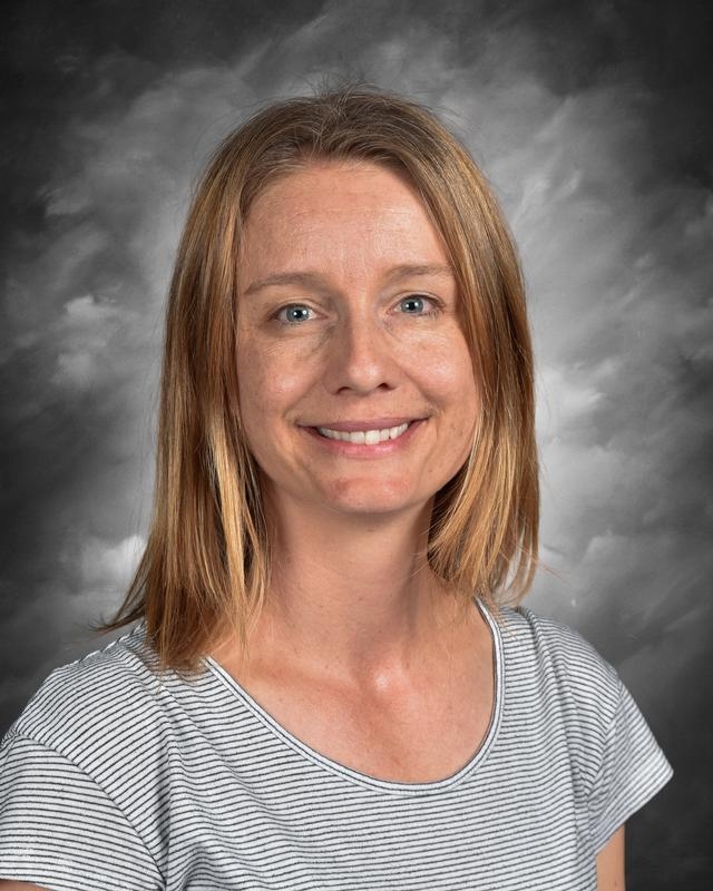 A woman with shoulder-length blonde hair smiles, wearing a striped shirt, against a gray background.