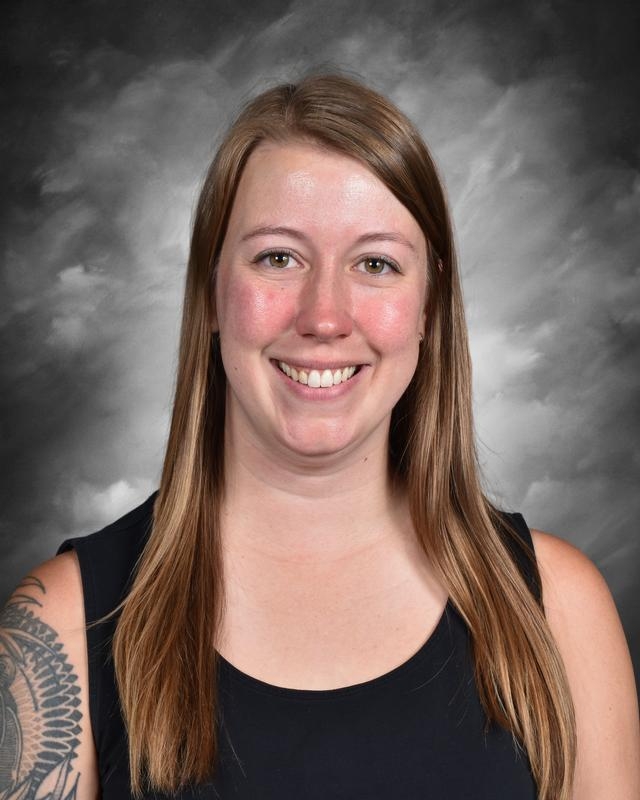 A woman with long brown hair smiles in front of a gray background, wearing a black sleeveless top.