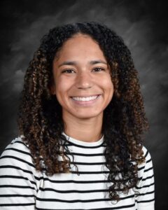 Smiling young woman with curly hair, wearing a striped shirt, against a dark background.