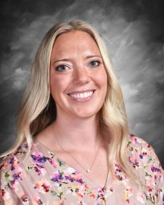Smiling woman with long blonde hair wearing a floral blouse against a cloudy gray background.