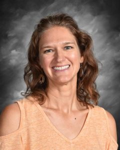 A woman with curly brown hair smiles, wearing a peach top and earrings, against a gray background.