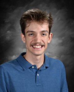 Smiling young man with short brown hair and a mustache, wearing a blue polo shirt against a dark, cloudy background.