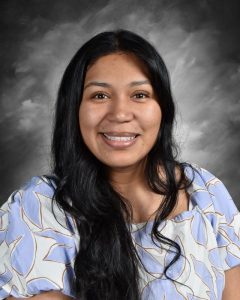 A smiling woman with long dark hair, wearing a light blue patterned blouse, is posed against a gray background.