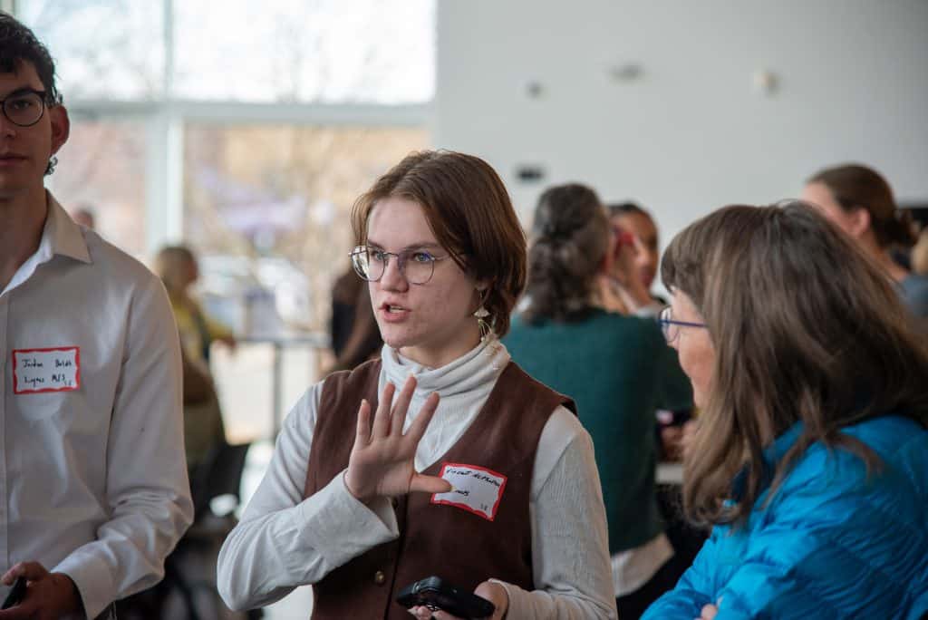 A young woman with glasses gestures while speaking in a crowded indoor setting, with others conversing around her.