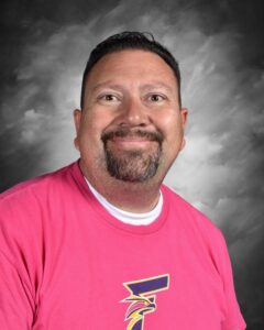 Smiling man with a beard wearing a pink shirt featuring a logo, set against a gray background.