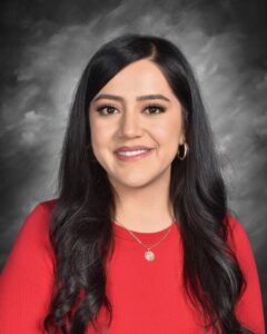 Smiling woman with long dark hair, wearing a red top and gold jewelry, against a dark background.