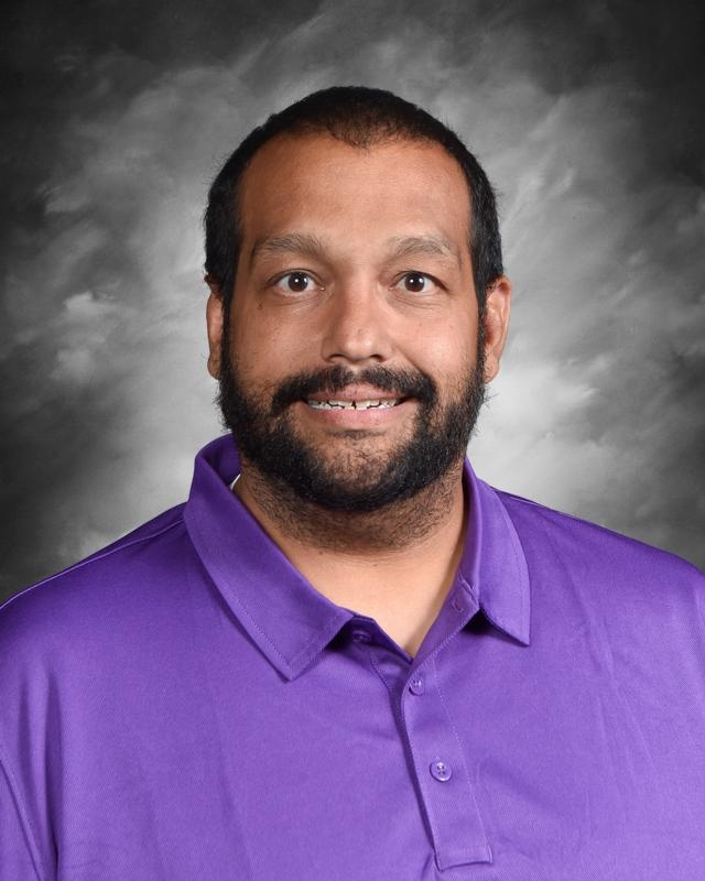 A smiling man with a beard wearing a purple polo shirt, set against a cloudy background.