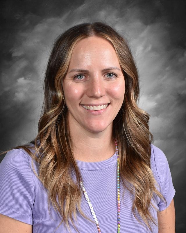 Smiling woman with long, wavy hair wearing a purple shirt and colorful necklace against a gray background.