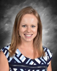 Smiling woman with long, straight hair wearing a patterned blue and white shirt, set against a gray background.