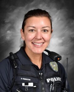 Smiling police officer in uniform with a badge and radio, set against a gray background.