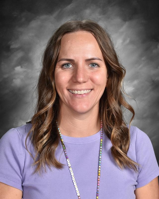 Smiling woman with wavy hair wearing a purple top and colorful beaded necklace, set against a gray background.