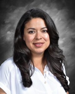 A woman with long, dark hair smiles, wearing a white blouse, against a softly blurred gray background.