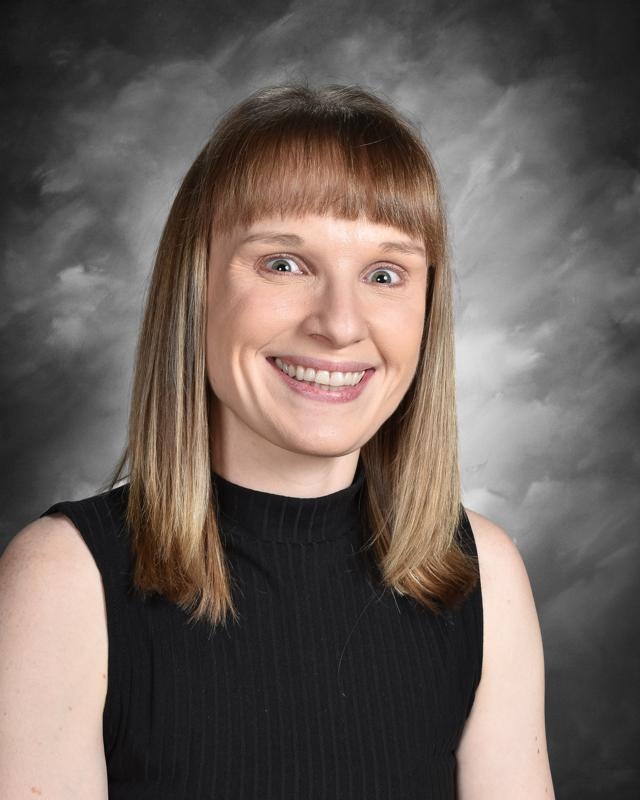 Smiling woman with straight, shoulder-length hair and bangs, wearing a sleeveless black top against a cloudy backdrop.