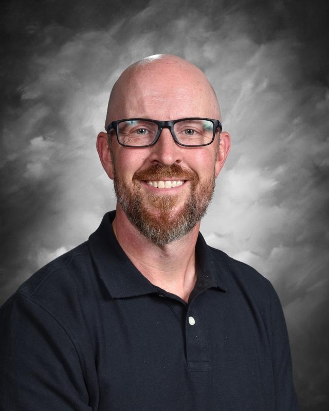 Bald man with glasses and a beard, wearing a dark polo shirt, smiling against a cloudy gray backdrop.